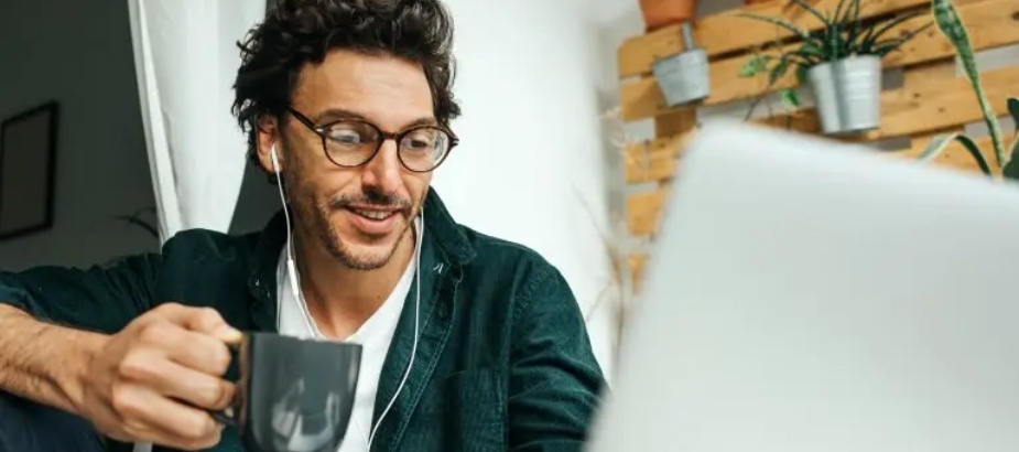 a man wearing glasses holding a mug and looking at his laptop